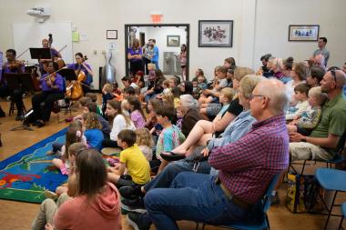 A portion of the SC Philharmonic Orchestra is visible while a large crowd watches them.