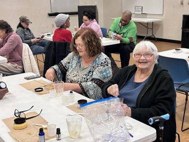 An elderly woman smiles at the camera while around her, several other people work on creating sugar scrubs.
